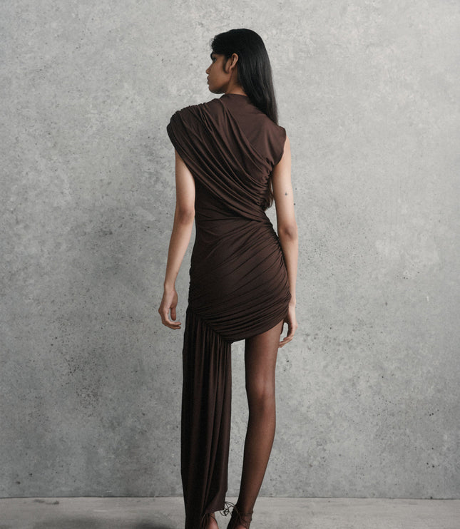 Rear view of a woman wearing the brown ruched asymmetrical mini maxi dress with sheer black tights and high heels.