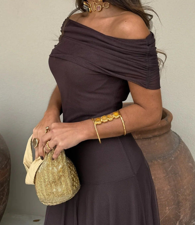Side profile of a women's chocolate brown off-the-shoulder mesh maxi dress, ruched sleeves, with gold accessories.