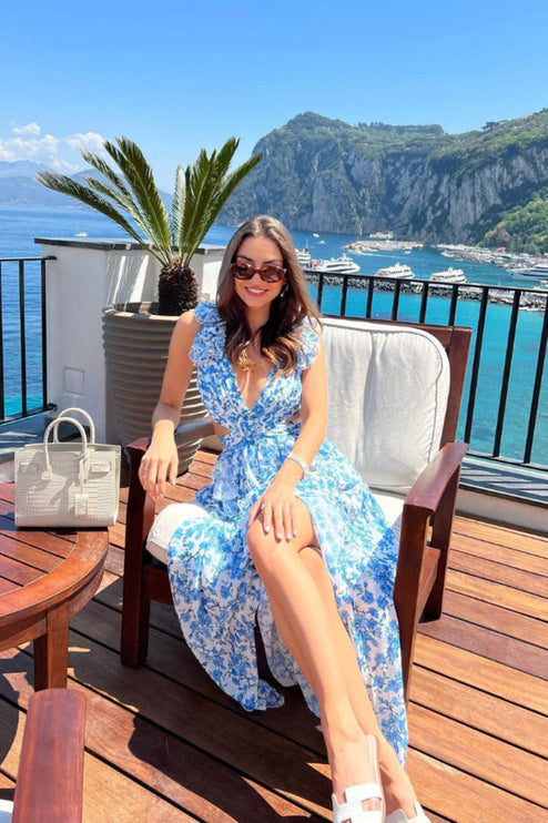 Women's blue and white floral print maxi dress with ruffles and V-neck, worn casually on a sunny balcony overlooking the sea.