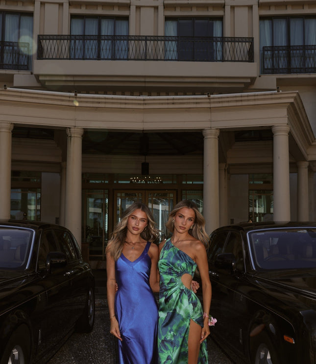 Two women wearing elegant maxi dresses, one in a blue slip style, the other in a green floral one shoulder cut out style, standing between black luxury cars in front of a grand building.