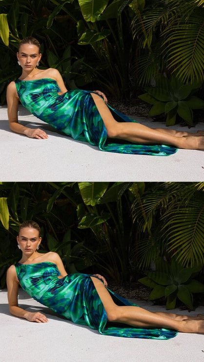 Women's one-shoulder floral maxi dress in vibrant green and blue, featuring a side cut-out and high slit, on a model lounging outdoors.