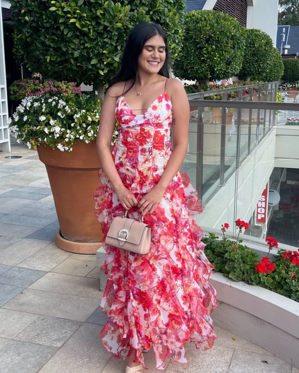 Smiling woman wearing a floral print ruffled spaghetti strap maxi dress, holding a beige handbag outdoors.