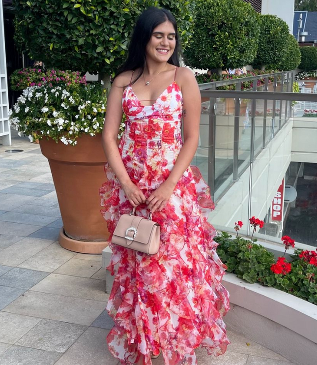 Smiling woman wearing a floral print ruffled spaghetti strap maxi dress, holding a beige handbag outdoors.