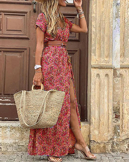 Women's red and pink paisley floral print short sleeve maxi dress with a brown belt and side slit, holding a straw bag.