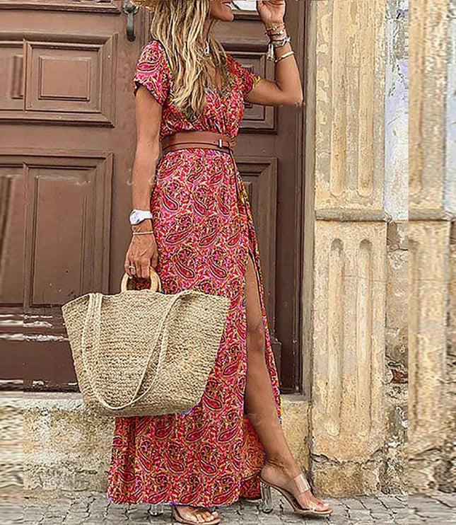 Women's red and pink paisley floral print short sleeve maxi dress with a brown belt and side slit, holding a straw bag.