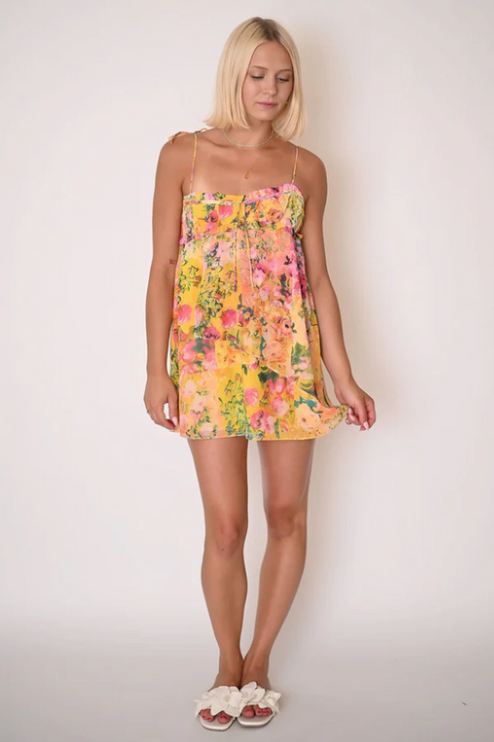 Full body shot of a woman wearing a yellow floral print tie shoulder mini dress with delicate spaghetti straps.