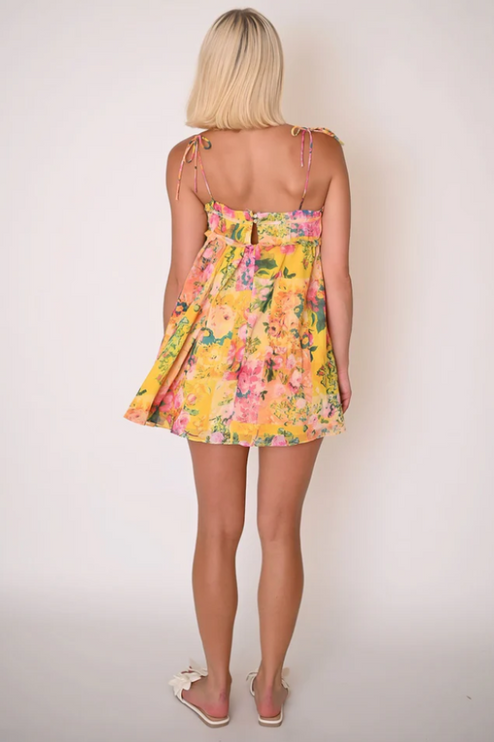 Back view of a woman wearing the yellow floral print tie shoulder mini dress, highlighting the flowy skirt.