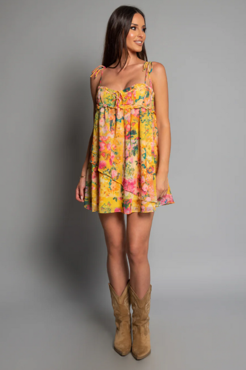 Full length view of a women's yellow floral print mini dress with tie shoulders, paired with cowboy boots.