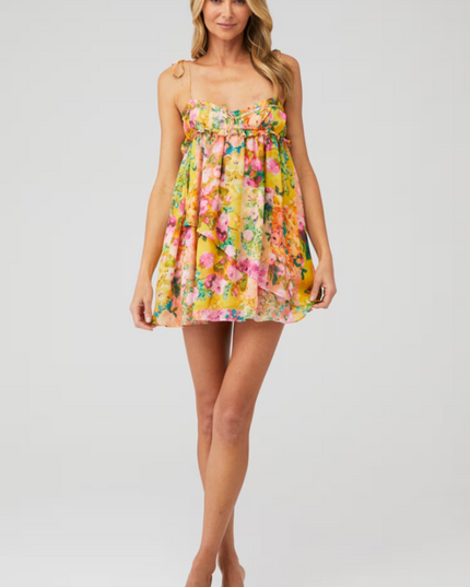 Front view of a woman posing in a bright yellow floral print tie shoulder mini dress with ruffled details.