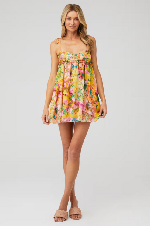 Front view of a woman posing in a bright yellow floral print tie shoulder mini dress with ruffled details.