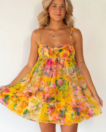 Front view of a blonde woman wearing a vibrant yellow floral print tie shoulder mini dress.