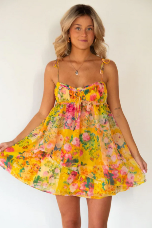 Front view of a blonde woman wearing a vibrant yellow floral print tie shoulder mini dress.