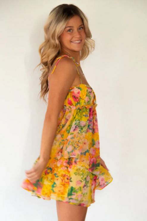 Side view of a blonde woman twirling in a yellow floral print tie shoulder mini dress.