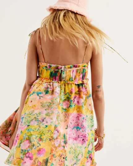 Back view of a women's yellow floral print tie shoulder mini dress with a pink bucket hat.
