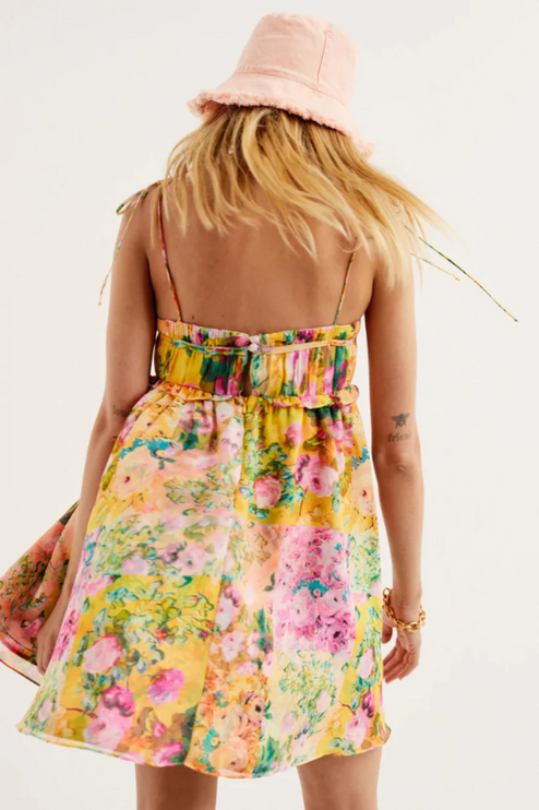 Back view of a women's yellow floral print tie shoulder mini dress with a pink bucket hat.