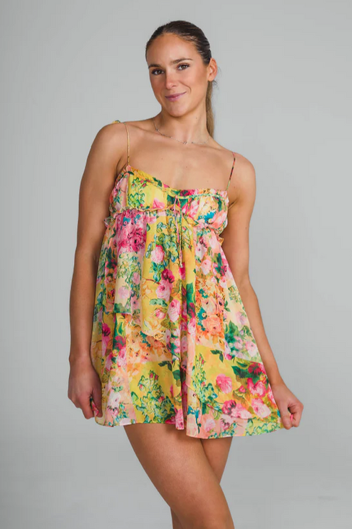 Front view of a woman wearing a yellow floral print tie shoulder mini dress, smiling at the camera.