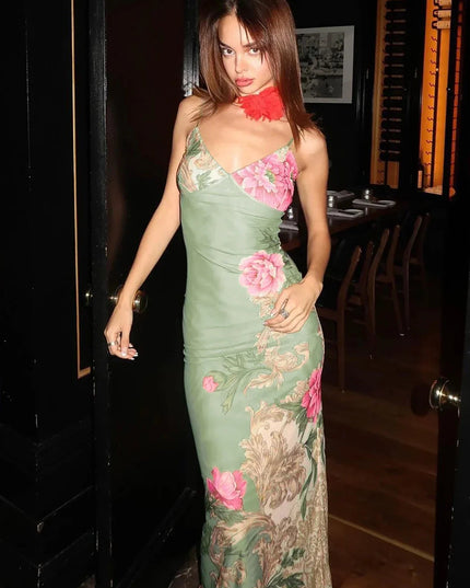 Full-length women's floral print V-neck maxi dress in sage green with large pink flowers and ornate scrollwork, shown in a doorway.