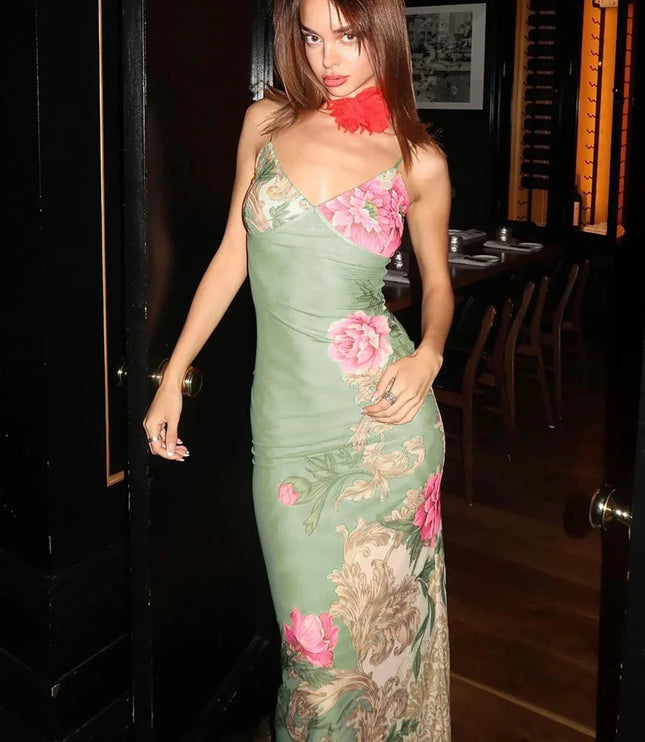Full-length women's floral print V-neck maxi dress in sage green with large pink flowers and ornate scrollwork, shown in a doorway.