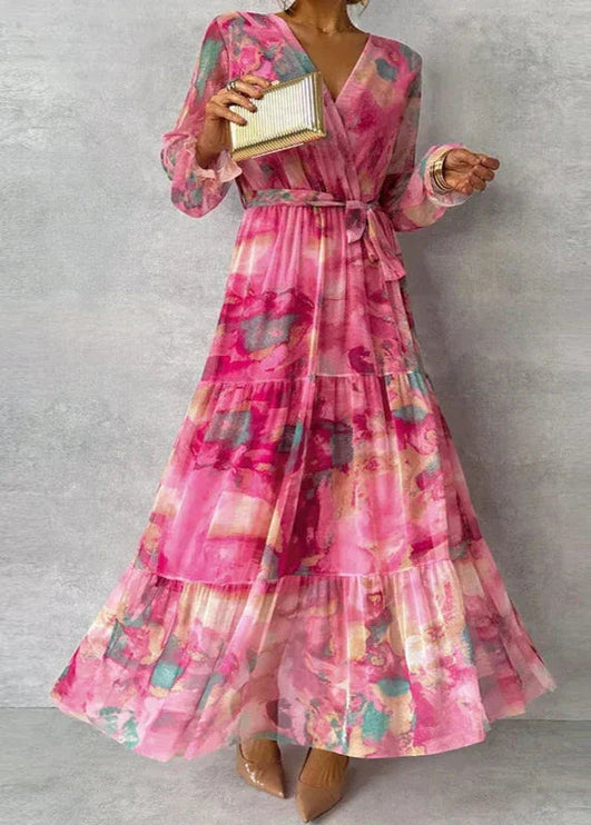 Women's pink floral printed V-neck long sleeve maxi dress with tiered skirt and tie waist, holding a gold clutch.