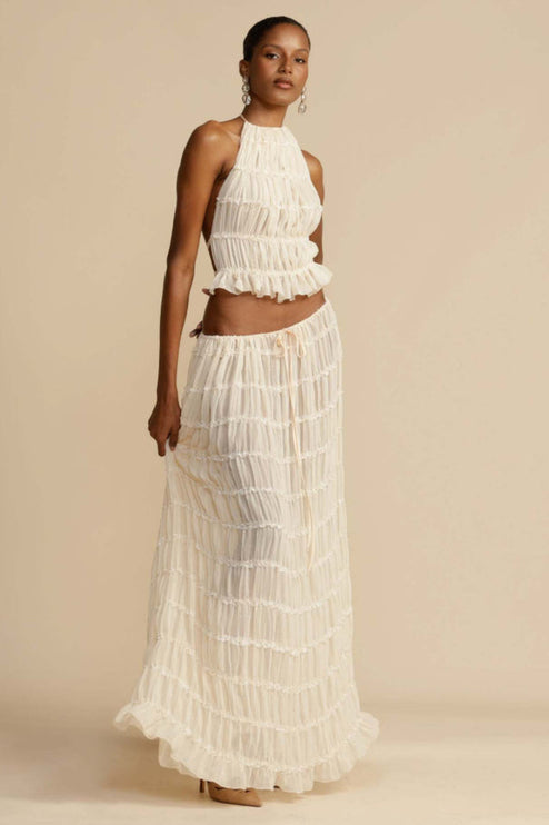 Front view of the women's ivory halter ruffle trim crop top and maxi skirt set, with the model holding the skirt fabric.