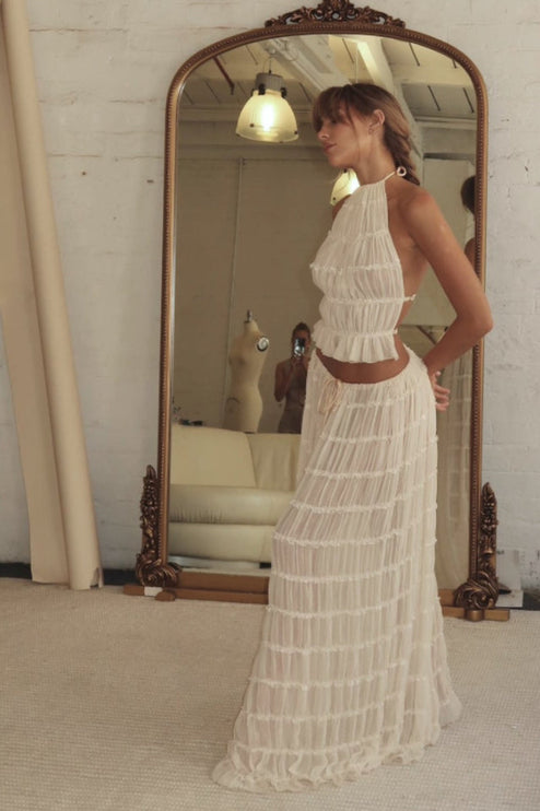 Side profile of a woman wearing the ivory halter ruffle trim crop top and maxi skirt set, reflected in a vintage mirror.