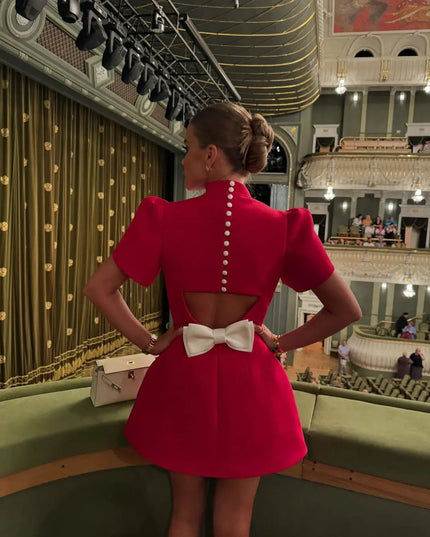 Back view of a red high neck mini dress with a keyhole cutout and white bow, shown in an elegant theater setting.