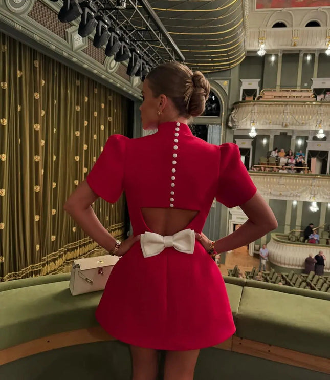 Back view of a red high neck mini dress with a keyhole cutout and white bow, shown in an elegant theater setting.