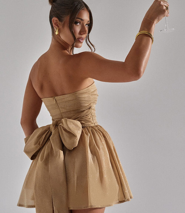 Back view of a woman in a gold strapless party dress, holding a champagne glass, highlighting the bow.