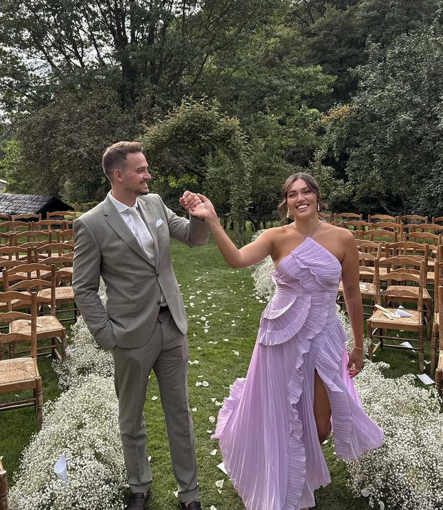 Women's lavender one-shoulder ruffle midi dress with a high slit, worn at an outdoor wedding with a man in a sage green suit.