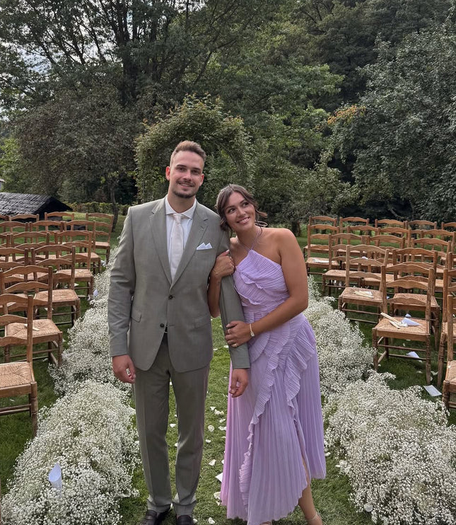 Lavender one-shoulder ruffle midi dress for women, paired with a man in a light green suit, posing outdoors at a wedding.