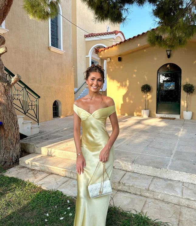 Women's light yellow off-shoulder satin maxi dress with a sweetheart neckline, styled with a white quilted handbag, standing outside a building.