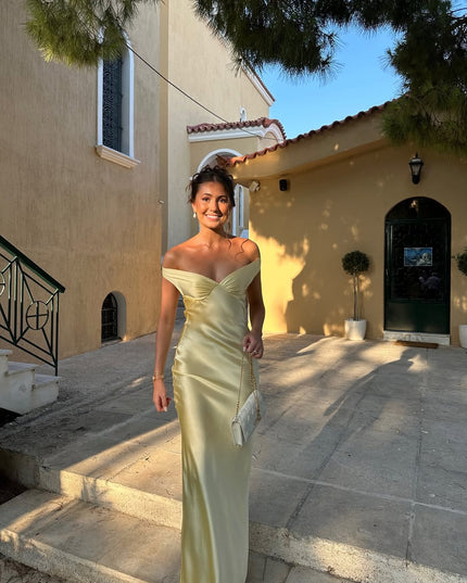 Smiling woman in a light yellow off-shoulder backless satin maxi dress, showcasing the dress's flow and elegant fit in an outdoor setting.