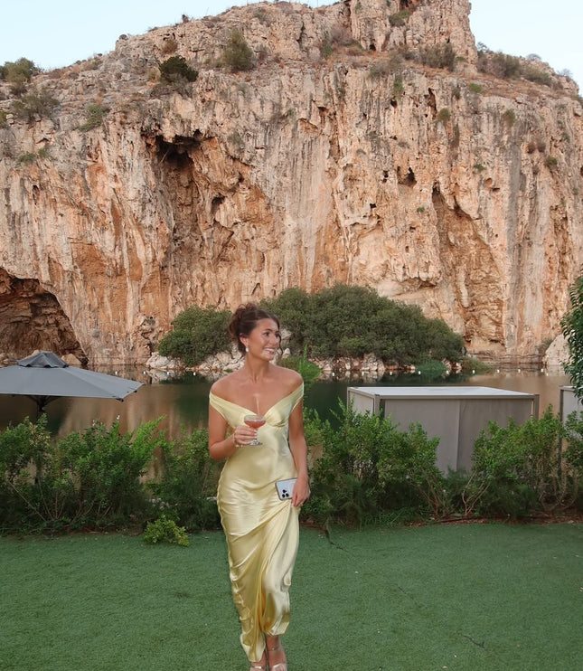 Women's light yellow off-shoulder satin maxi dress with a flowing skirt, worn outdoors with a drink in hand, against a scenic rock and water backdrop.
