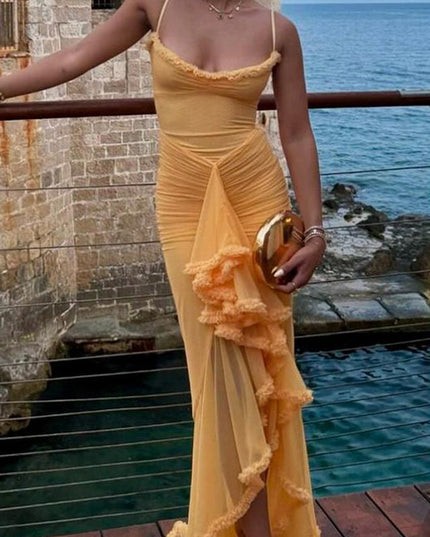 Women's yellow prom ruffled front maxi dress worn outdoors by the water, holding a gold clutch, with layered necklaces.