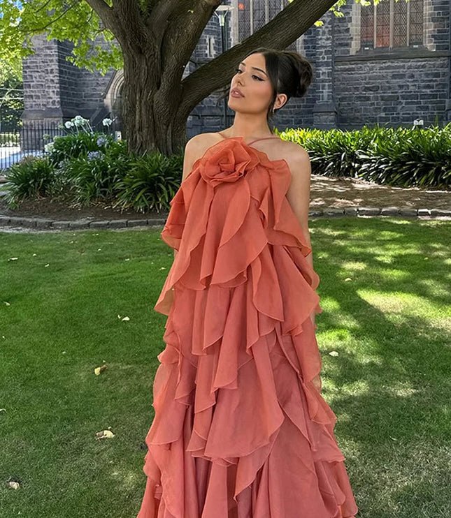 Woman posing outdoors in a coral strapless ruffle layered maxi dress, featuring a prominent floral detail on the bust, against a green lawn and tree.