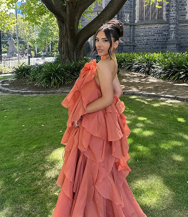Back view of a woman wearing a coral strapless ruffle layered maxi dress, showcasing the tiered skirt and a subtle back strap detail, in a garden setting.