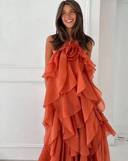 Front view of a smiling woman wearing a strapless coral prom maxi dress with a large ruffled flower detail on the bust and tiered ruffles cascading down the skirt.