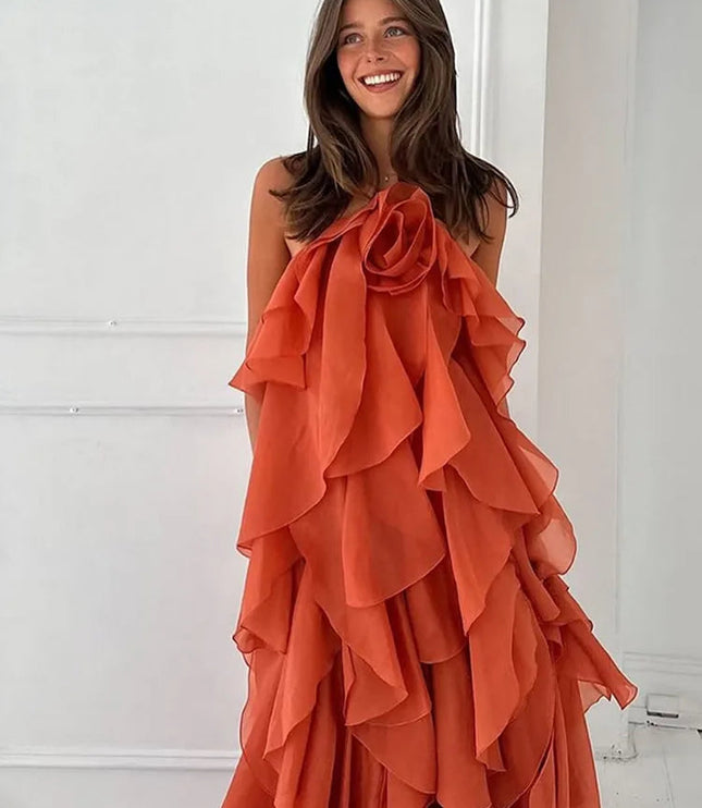 Front view of a smiling woman wearing a strapless coral prom maxi dress with a large ruffled flower detail on the bust and tiered ruffles cascading down the skirt.