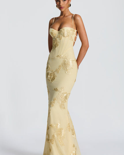 Side view of a woman wearing a light yellow floral lace-up maxi dress, featuring gold embroidered flowers and a silhouette.