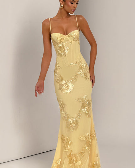 Woman modeling a light yellow floral lace-up maxi dress with gold embroidered flowers, showing the dress's full length and design.