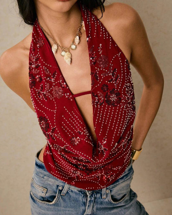 Front view of a women's red sequin halter deep V neck top, draped over light wash jeans, with a shell necklace.