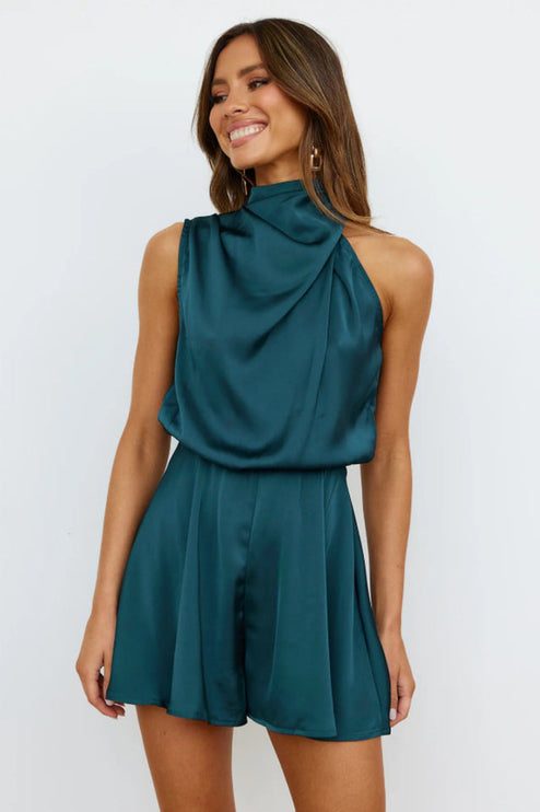 Front view of a woman wearing a sleeveless high neck satin romper in a deep teal color, featuring a draped neckline and elasticized waist.