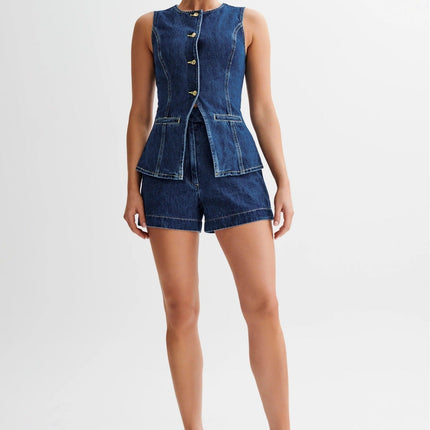 Sleeveless single breasted denim shorts set in dark wash, featuring gold buttons and a peplum waist, paired with white heels.