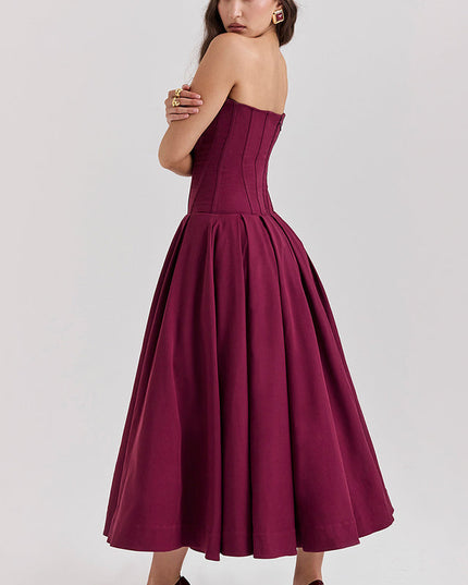 Rear side view of a woman in a deep burgundy strapless boned bodice pleated midi dress, arms crossed.
