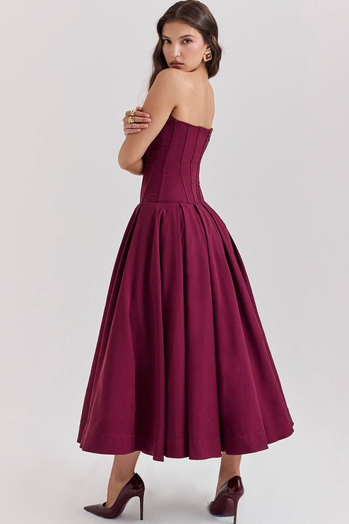 Rear side view of a woman in a deep burgundy strapless boned bodice pleated midi dress, arms crossed.