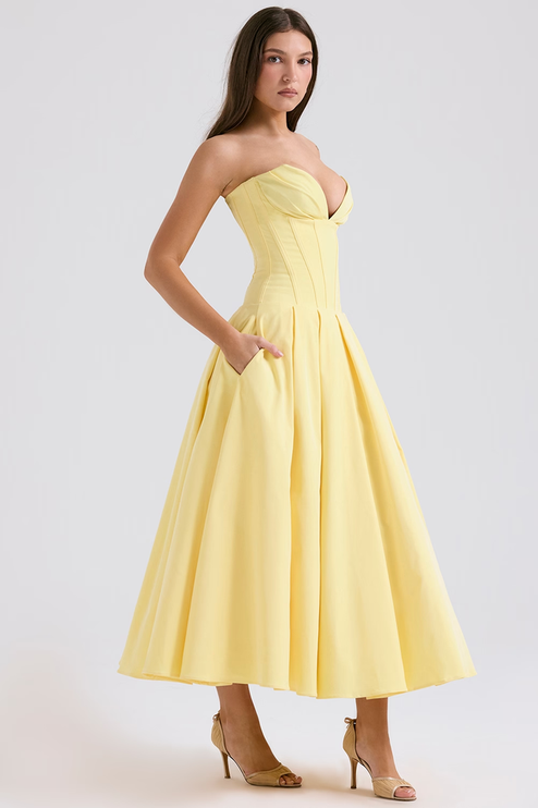 Side profile of a yellow strapless boned bodice pleated midi dress with pockets, paired with nude heels.
