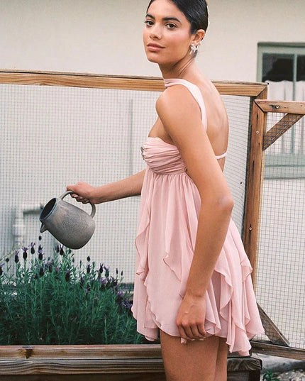 Side profile of a woman wearing a light pink ruffle halter mini dress, watering plants in a garden.