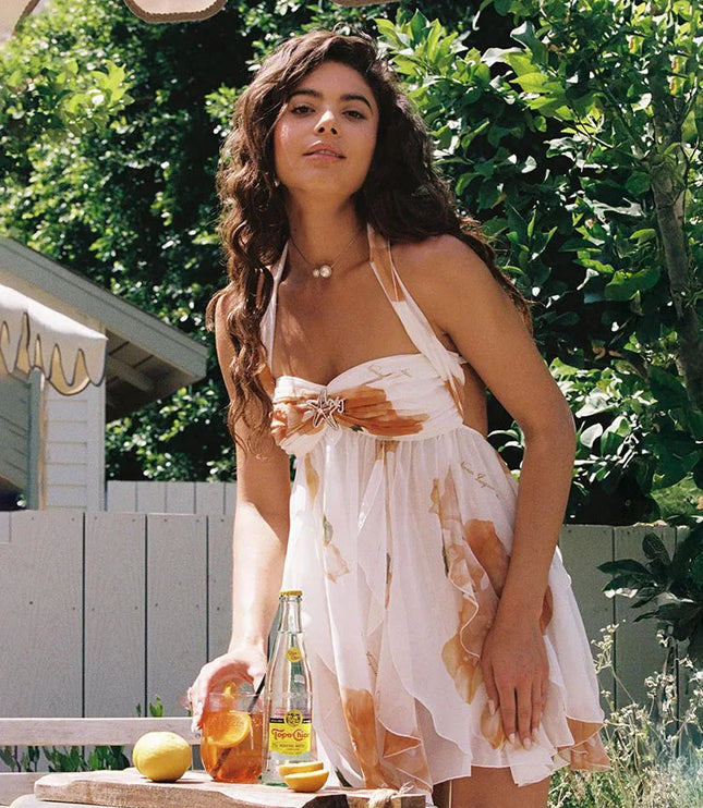 Woman wearing a white summer floral ruffle halter mini dress with orange flowers, enjoying a drink outdoors.