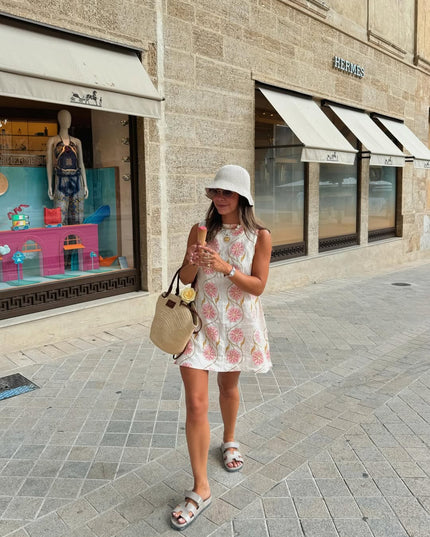 Woman walking on a street, wearing a white summer floral mini dress, white bucket hat, and light grey slides.