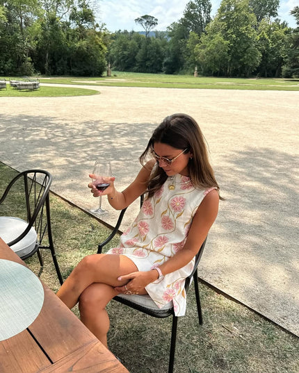 Woman seated outdoors in a black chair, wearing the white summer floral sleeveless mini dress and holding a glass of wine.